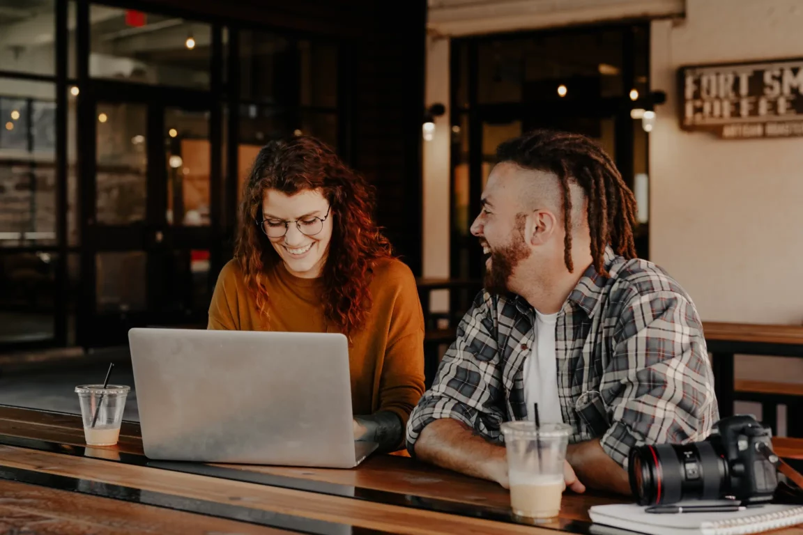 Two friends at a cafe, woman with curly hair using a laptop, man with dreadlocks laughing.