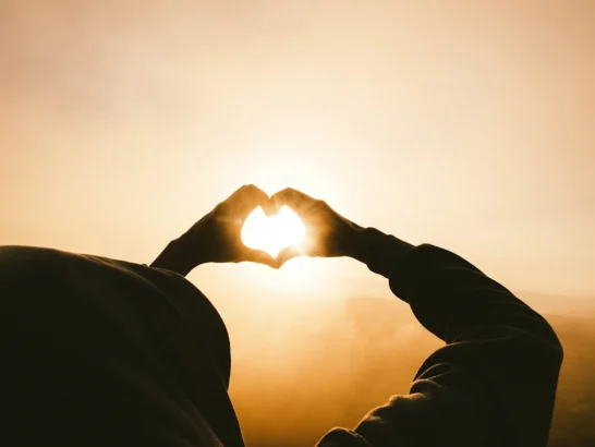 Silhouette of hands forming a heart shape around the bright sun during a golden sunset.